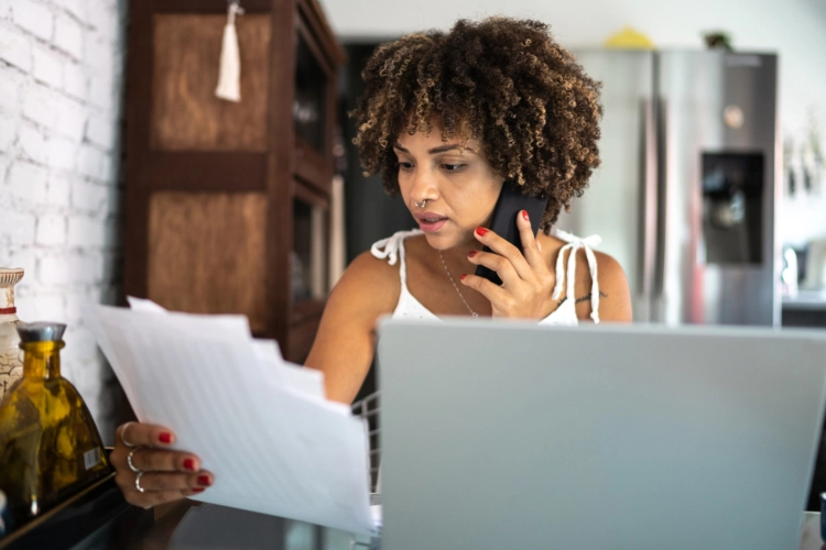 A woman with curly hair sits at a table with a laptop, holding a stack of documents and talking on a mobile phone.