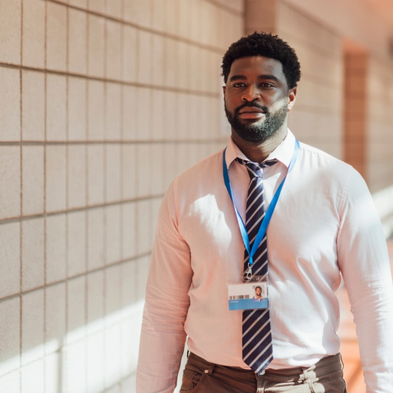 A man wearing a light pink button-down shirt, a striped tie, and an ID badge walks down a tiled hallway.