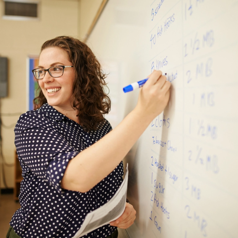 A smiling woman wearing glasses and a polka-dot shirt writes a list of items on a whiteboard with a blue marker.