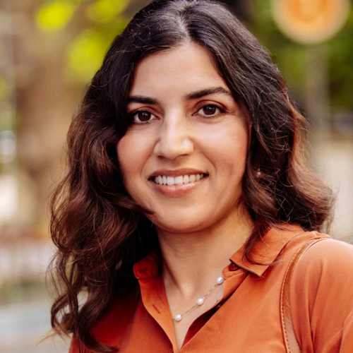 A smiling woman with dark, wavy hair and an orange shirt stands outdoors, looking directly at the camera.