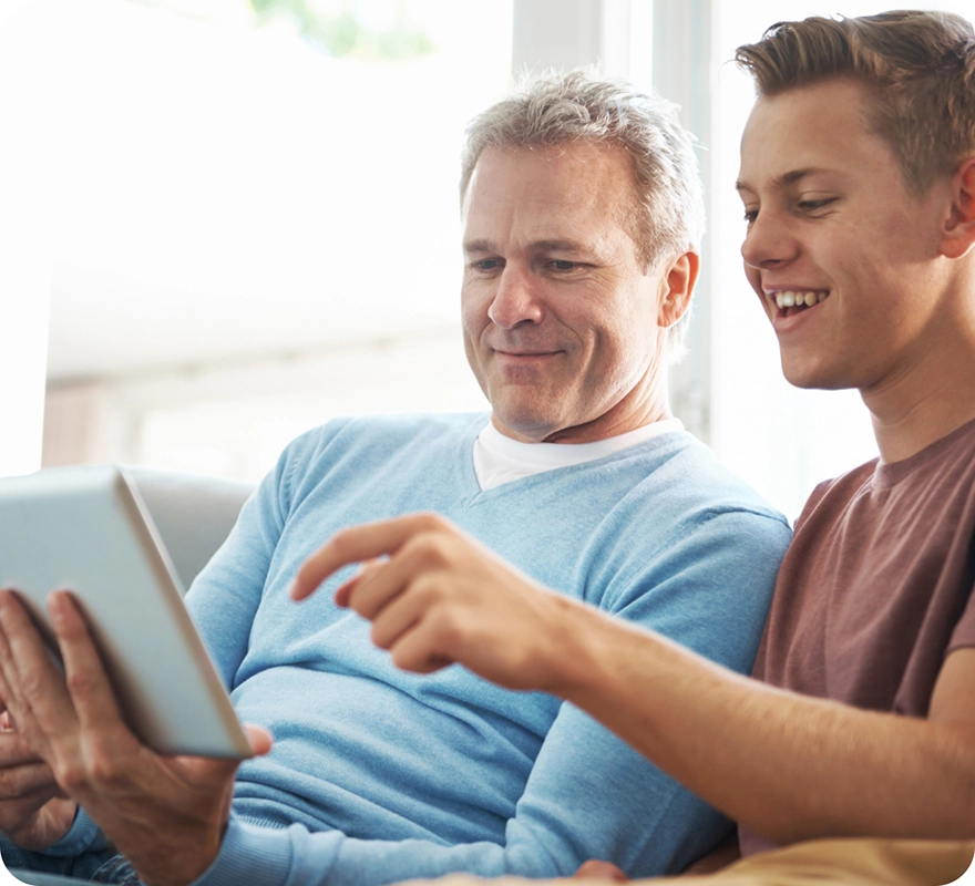A father and son sitting on the couch and reviewing medical information on an iPad.