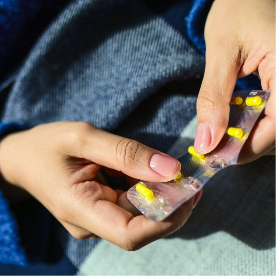A close-up of a person's hands breaking open a blister pack of yellow pills. Overlaid on the image is an app notification confirming "Pharmacy confirmed" with operating hours of "7:00 AM - 9:00 PM."